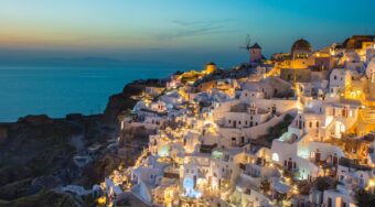 View from Oia, with lit up buildings on the cliff edge looking out towards a blue sea at dusk.