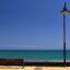 A wooden seat next to a lamp on a yellow paved footpath with a blue sky and ocean in the background.