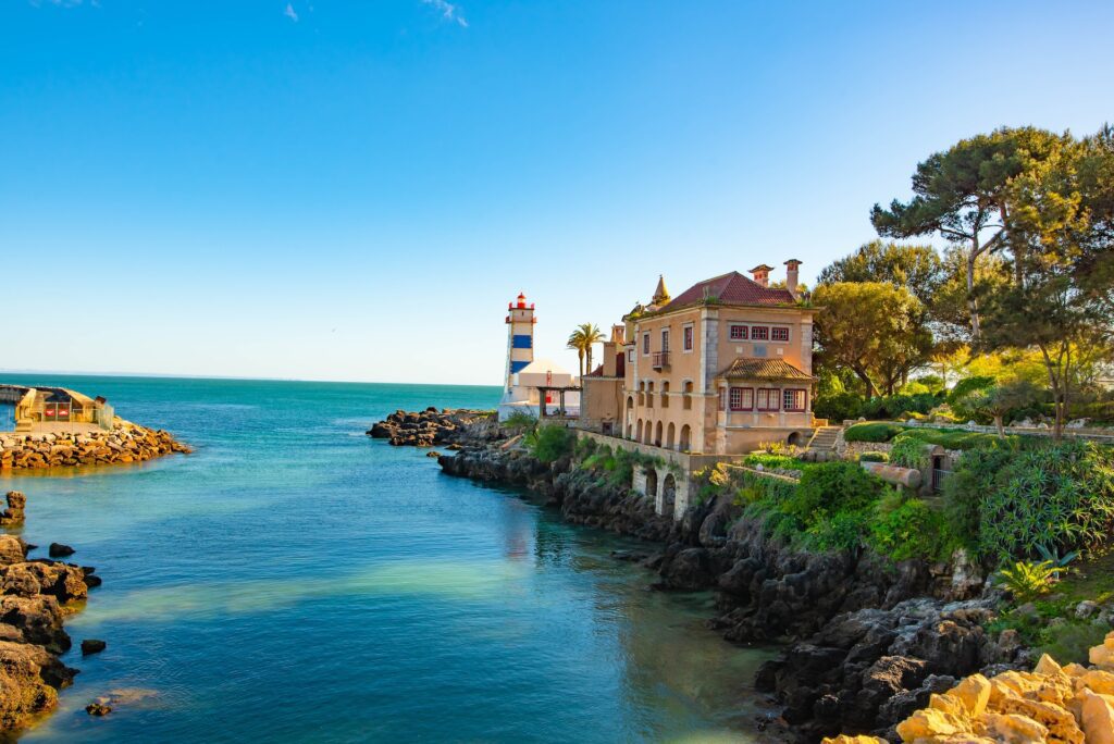 View of blue waters off the coast of Cascais, surrounded by rocks and palms, with a historical building perched by the water. Take a taxi from Lisbon Airport to Cascais for a relaxing start to your trip.
