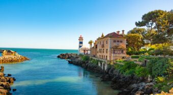 View of blue waters off the coast of Cascais, surrounded by rocks and palms, with a historical building perched by the water. Take a taxi from Lisbon Airport to Cascais for a relaxing start to your trip.