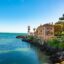 View of blue waters off the coast of Cascais, surrounded by rocks and palms, with a historical building perched by the water. Take a taxi from Lisbon Airport to Cascais for a relaxing start to your trip.