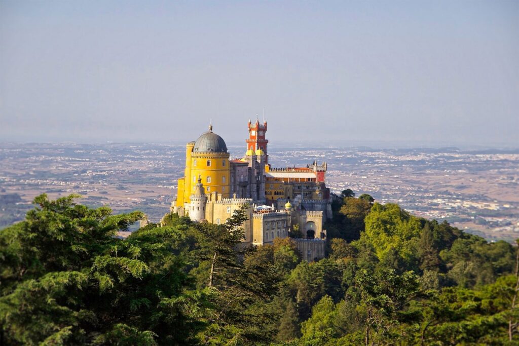 View of a colourful castle on a hilltop in Sintra, surrounded by lush greenery and blue skies above. With a taxi from lisbon airport to sintra you'll get to see it as soon as you land.