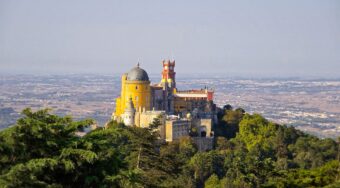 View of a colourful castle on a hilltop in Sintra, surrounded by lush greenery and blue skies above. With a taxi from lisbon airport to sintra you'll get to see it as soon as you land.