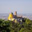 View of a colourful castle on a hilltop in Sintra, surrounded by lush greenery and blue skies above. With a taxi from lisbon airport to sintra you'll get to see it as soon as you land.
