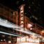 A stylish nighttime photo of Chicago's downtown. A bright lit sign reading "Chicago" is vertical in the center of the photo, likely for a Broadway theatre.