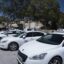 A line of white Malta taxis parked under the shade of a tree.