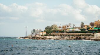 Scenic daytime view of the rocky coastline and waterfront near St Julian's, Malta, with the sea in the foreground and buildings along the shore, capturing the coastal atmosphere travelers enjoy when heading to St Julian's from Malta Airport.