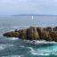 A rocky jetty surrounded by blue sea and waves under a cloudy sky with a sailboat in the distance.