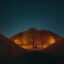 Autumn trees beneath a night sky in between the crux of two symmetrical hills that are lit up so they appear golden.