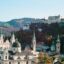 A cluster of white buildings with green and brown rooftops, some domed and others with tall spires, surrounded by trees and a mountain.