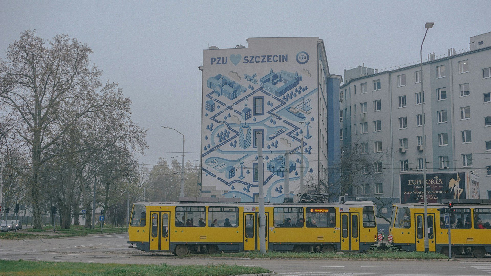 Three yellow trams parked in front of a white concrete building on a foggy morning, with blue graffiti-style art featured on the main face of the building.
