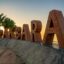 A large orange Pak Bara sign written with individual lettering placed on a concrete bollard on the Pak Bara pier with the ocean in the background and a couple of palm tree to the left of the sign.