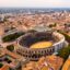 An aerial view of an ancient colosseum surrounded by a sprawling city with terracotta roofs.