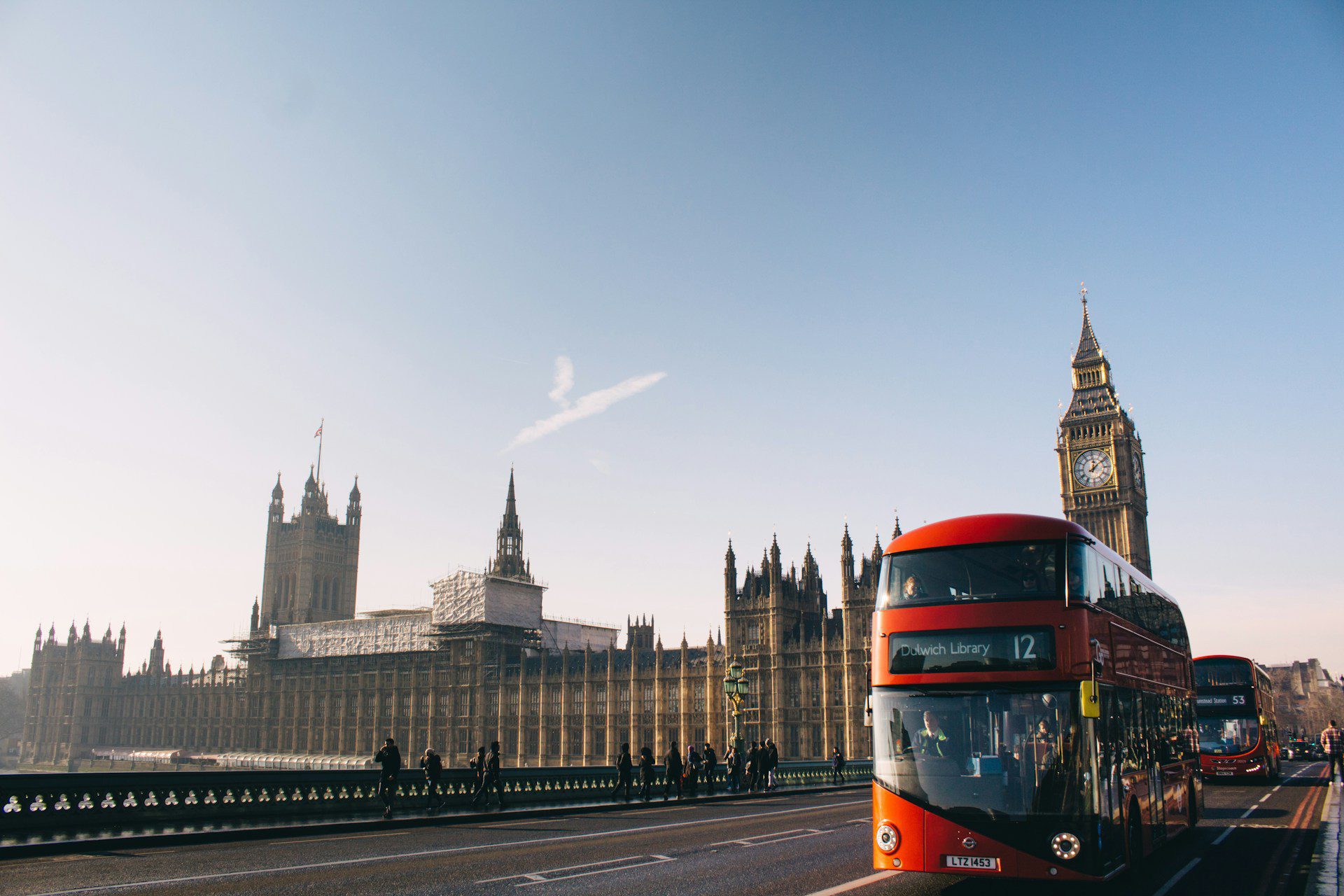 View of the houses of parliament and Big Ben from the street, with a red double-decker bus driving past, and blue skies above.