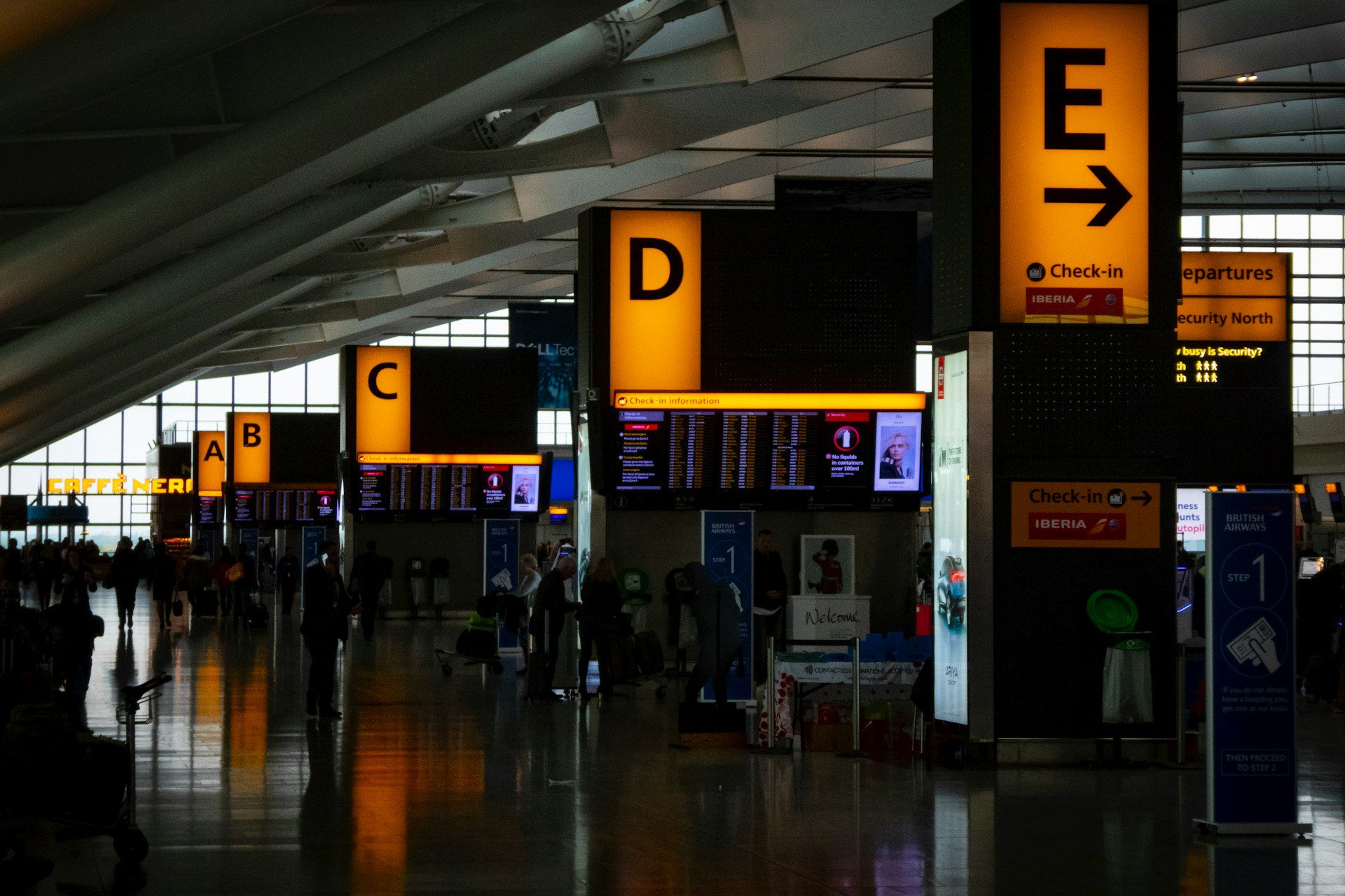 A photo of the check in desks inside Heathrow Airport terminal, showing orange glowing signs with letters on.