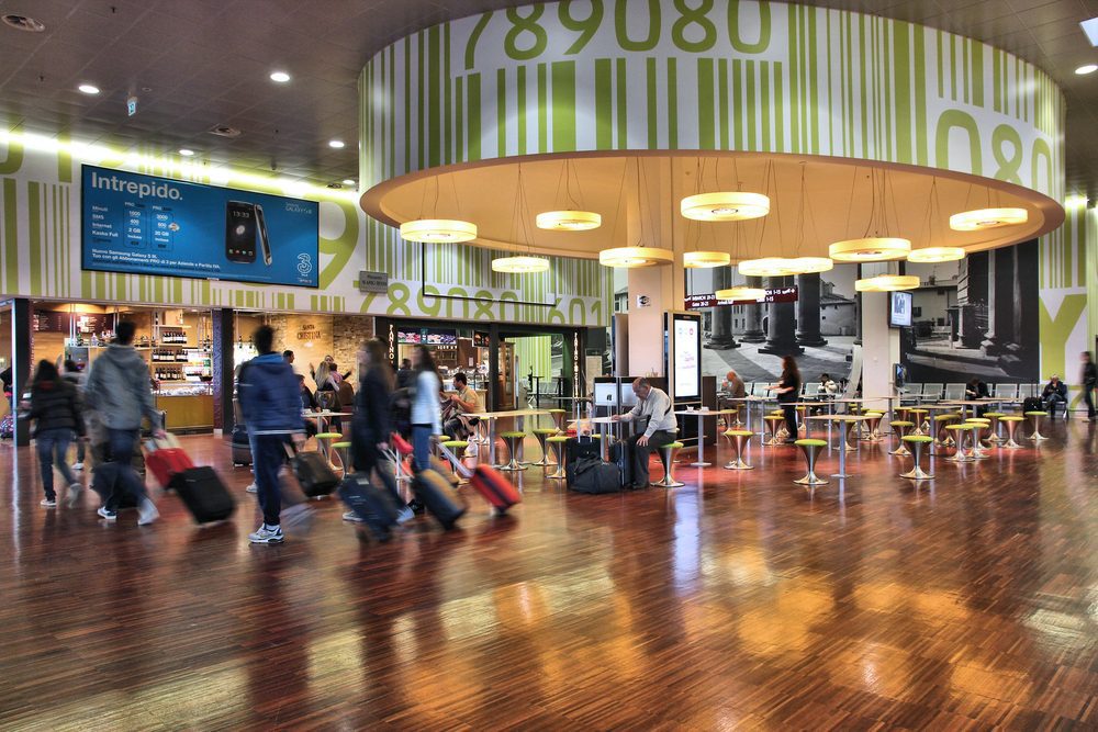 A blurred image of passengers passing through Bergamo Airport with polished floors and a central gathering area.