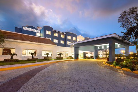 Evening view of a well-lit hotel entrance near Airport Cancun, featuring a curved driveway, modern exterior design, and landscaped greenery under a colorful sky.
