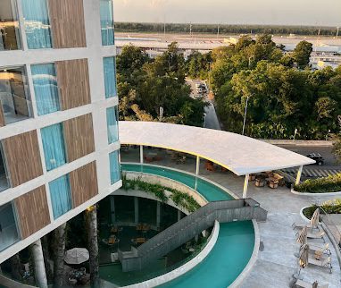 View of a modern hotel near Airport Cancun, featuring a circular pool with a footbridge, shaded outdoor seating, and nearby road access surrounded by trees.