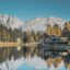 Mountains and tall trees line a large cabin reflecting on the water in Madonna di Campiglio.