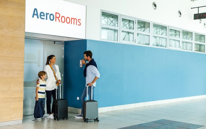 Exterior of AeroRooms Hotel in Terminal 1 at Prague Airport, with a white sign above the door reading AeroRooms, and people with luggage standing in front of the door.