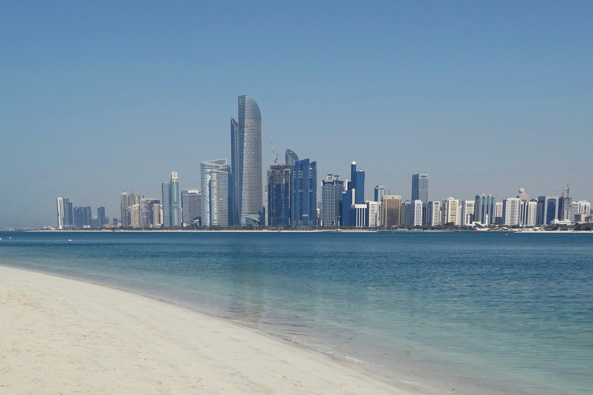 View of skyscrapers in central Abu Dhabi, with a blue body of water and white sands in front.