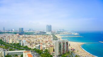 View of Barcelona City Beach, with skyscrapers on the left and a long stretch of sand meeting a blue sea on the right.