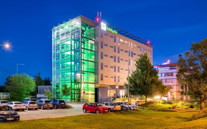 Exterior of the Holiday Inn at Prague Airport, with green light shining on the front of the building and dark skies above.