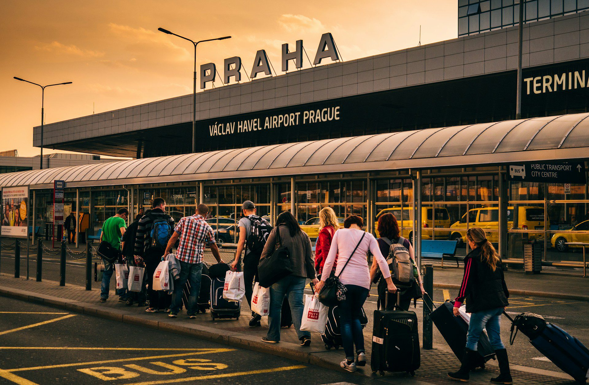 Exterior of Prague Airport, with a sign on the roof saying "Praha" and an orange sky above.