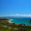 An aerial view of a snake-bend coastline with green pastures to the left of the sandy shore and bright blue ocean to the right.