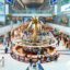 Busy interior of Dubai Airport DXB during daytime, featuring a large golden palm tree centerpiece, luxury shops like Tom Ford and Yves Saint Laurent, and travelers exploring the bustling duty-free area under a high arched ceiling.