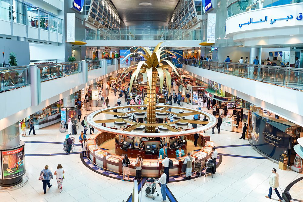 Busy interior of Dubai Airport DXB during daytime, featuring a large golden palm tree centerpiece, luxury shops like Tom Ford and Yves Saint Laurent, and travelers exploring the bustling duty-free area under a high arched ceiling.