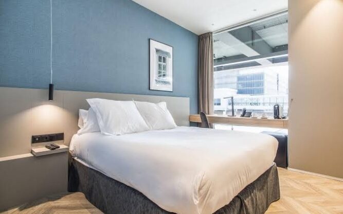 Hotel room with a double bed in the centre, a cornflower blue wall, and a window to the right.