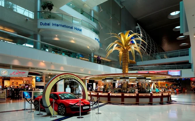Interior of Dubai International Airport featuring the Dubai Airport Hotel signage above the duty-free area, with luxury shops, a red sports car display, and a golden palm centerpiece under modern lighting.