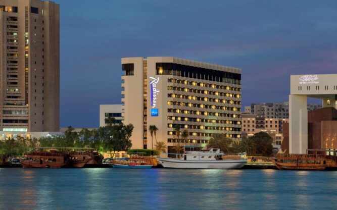 Waterfront view of Radisson Blu, a high-rise hotel near Dubai Airport, seen at dusk with traditional boats docked along Dubai Creek and city lights reflecting on the water.