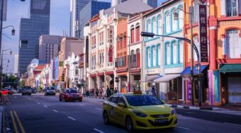 A yellow taxi and private cars driving down a street in Singapore, with colourful buildings either side.