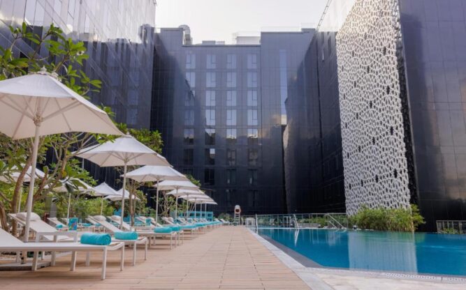 Outdoor pool area at a modern hotel near Dubai Airport, featuring lounge chairs with turquoise cushions, white umbrellas, and sleek black building facades surrounding the space.