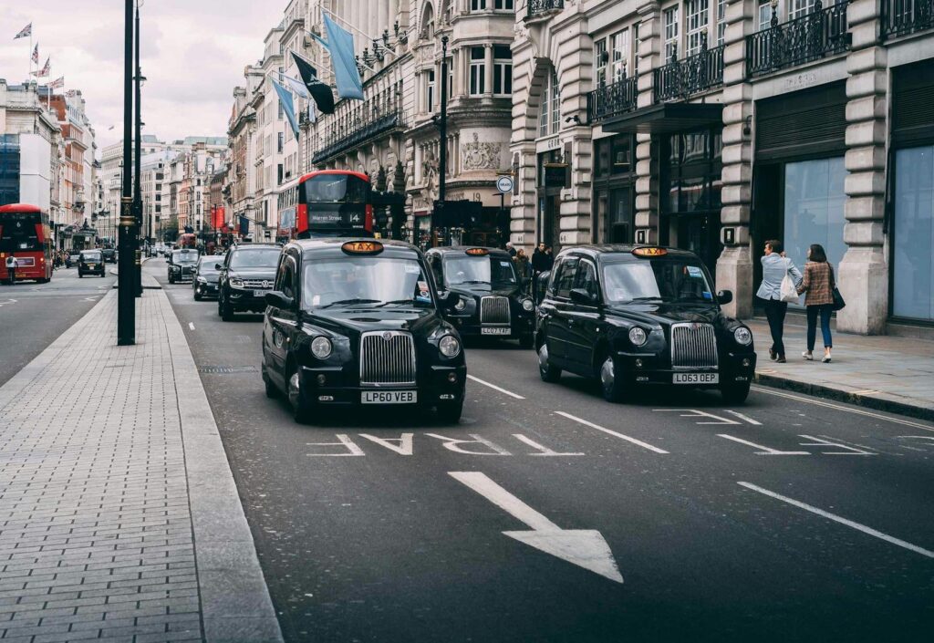 London Black Cabs driving in two lines along a London road, with a red double-decker bus in the background and buildings either side.