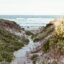 Wooden plank walkway between two greenery covered sand banks leading to the ocean.