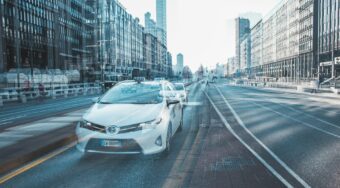 A blurry image of a white taxi driving on a road in central Milan, with buildings either side.