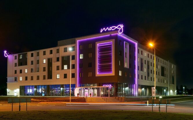 The Moxy Edinburgh Airport Hotel at night lit up with purple neon lights and signage.