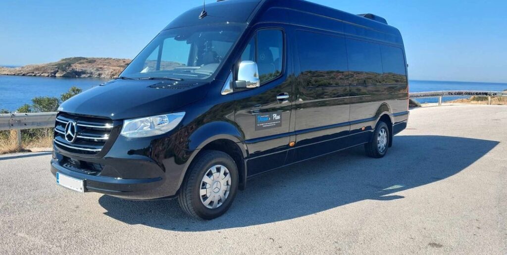 Side view of a black Athens Airport taxi minibus parked on the coast, with a body of water in the background.