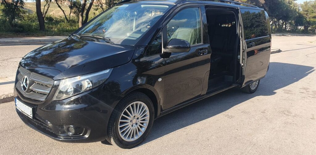 Side view of a black Athens Airport taxi minivan parked in a wooded area.