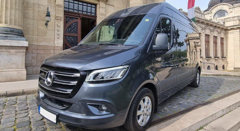 A black Budapest Airport taxi minibus parked in the street outside an old building.