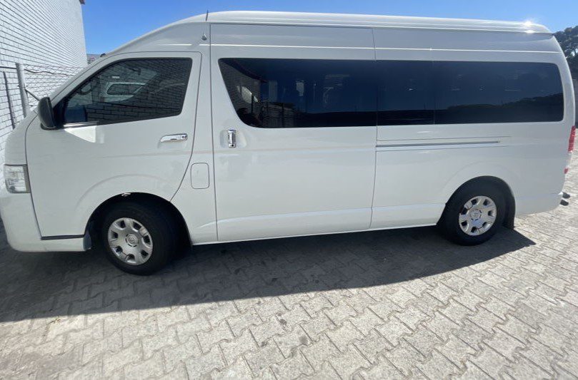 Taxi Cape Town Airport (Minibus) - Welcome Pickups Side view of a white Cape Town Airport taxi minibus parked outside a house, with blue skies above.