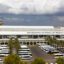 Panoramic view of Palma de Mallorca Airport showing the main terminal building with "Aeropuerto de Palma de Mallorca" signage, a bustling parking lot filled with tour buses and palm trees in the foreground, and the control tower in the distance beneath a dramatic, overcast sky.