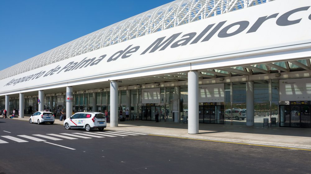 Exterior of Mallorca Airport on a clear day, showing the main terminal entrance with large glass windows, white signage, and taxis parked at the curb.