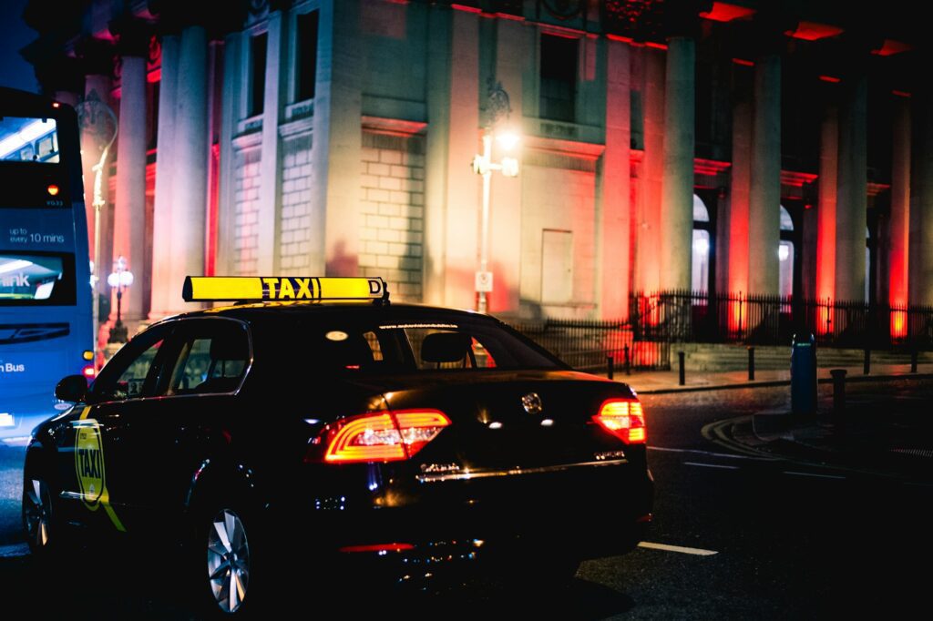 Image of a black taxi with a yellow taxi sign on the roof, in the streets at night.