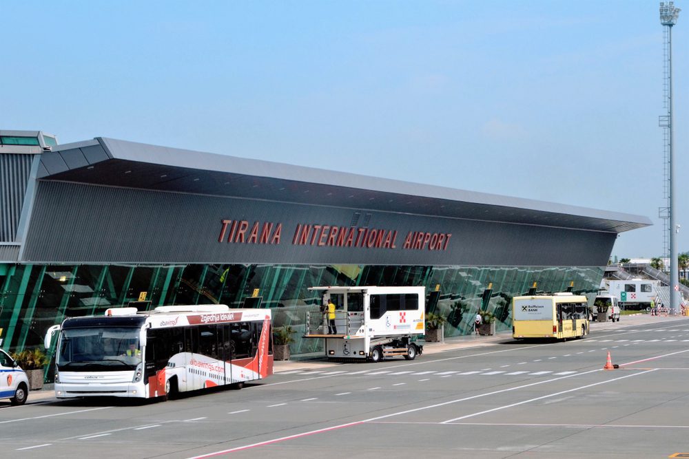 Front view of the main terminal at Tirana Airport, with buses parked outside and blue skies above.