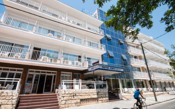 Daytime view of Hotel Amic Gala in Mallorca, featuring a modern multi-story facade with glass balconies, a blue glass entrance tower, and a cyclist passing on the quiet street in front.