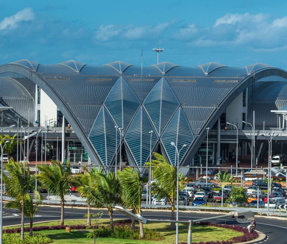The domed front entrance to Mauritius International Airport made of glass panels with a car park and palm trees in front.